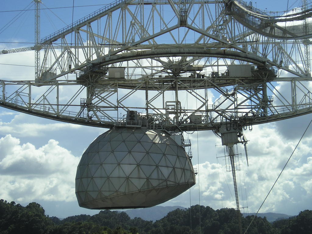 A suspended metal framework and large domed receiver above wooded hills beneath a blue sky with clouds.