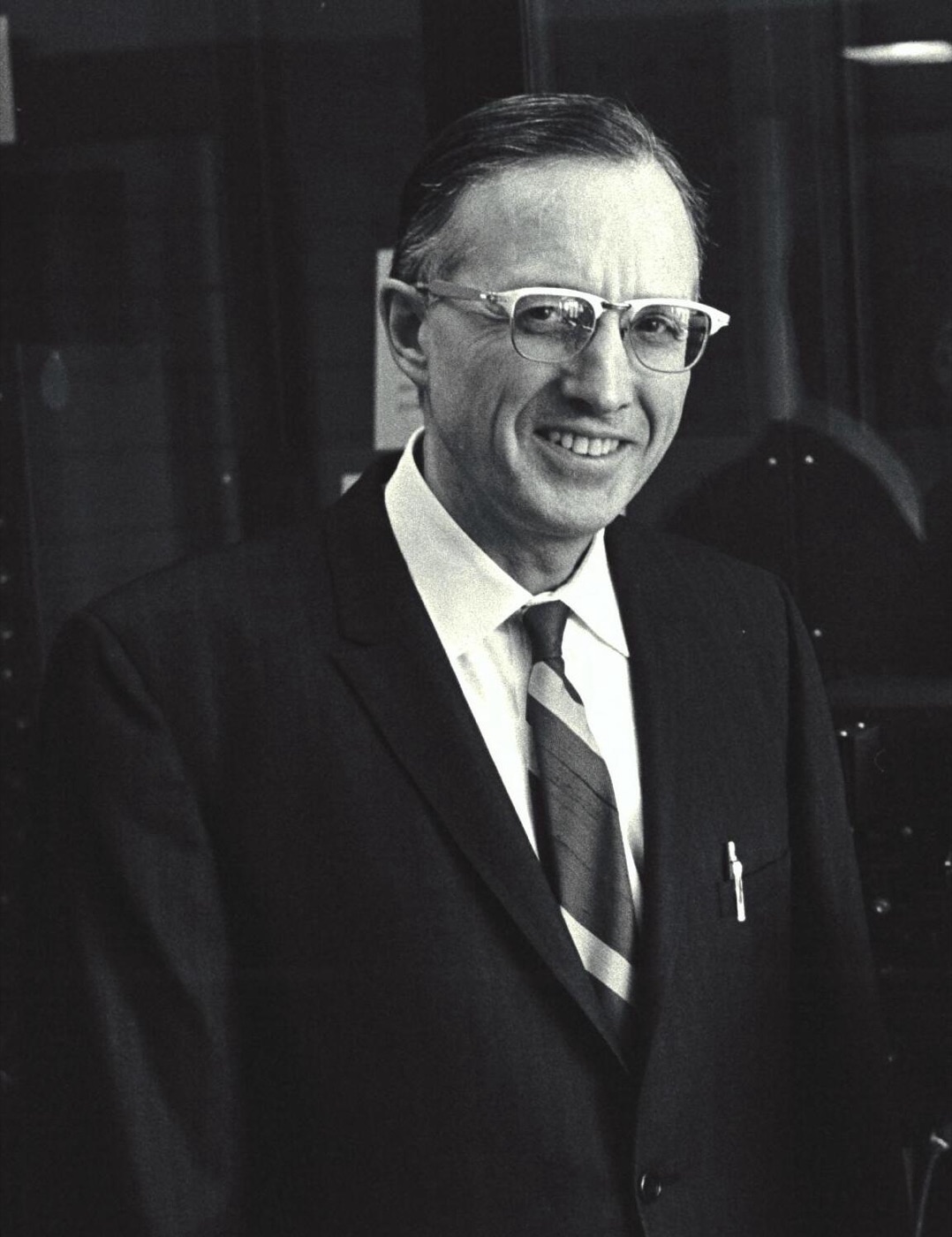 Smiling man in glasses, dark suit and striped tie photographed in front of dark equipment panels