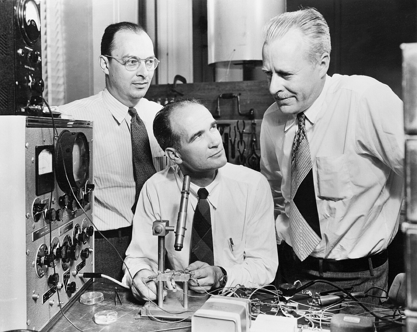 Three men in shirtsleeves around a laboratory bench with electronic test equipment; one sits adjusting a small apparatus