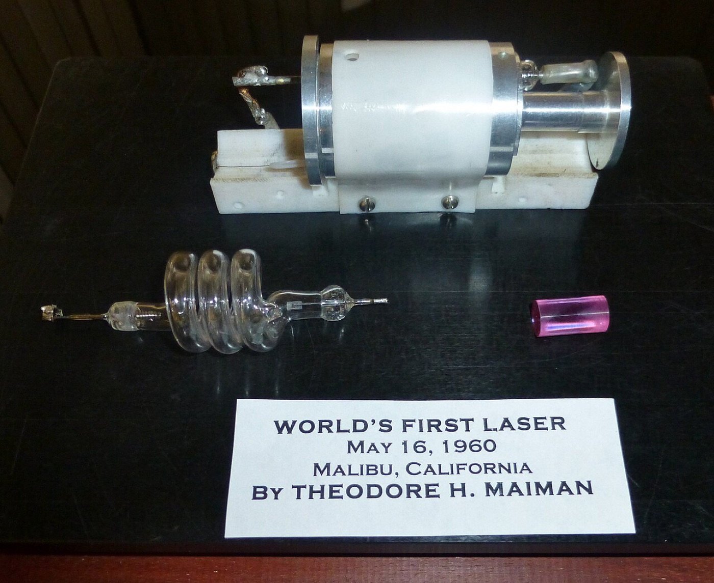 A ruby rod, a spiral glass flashlamp and a white cylindrical housing on a black surface above a printed label reading World's first laser