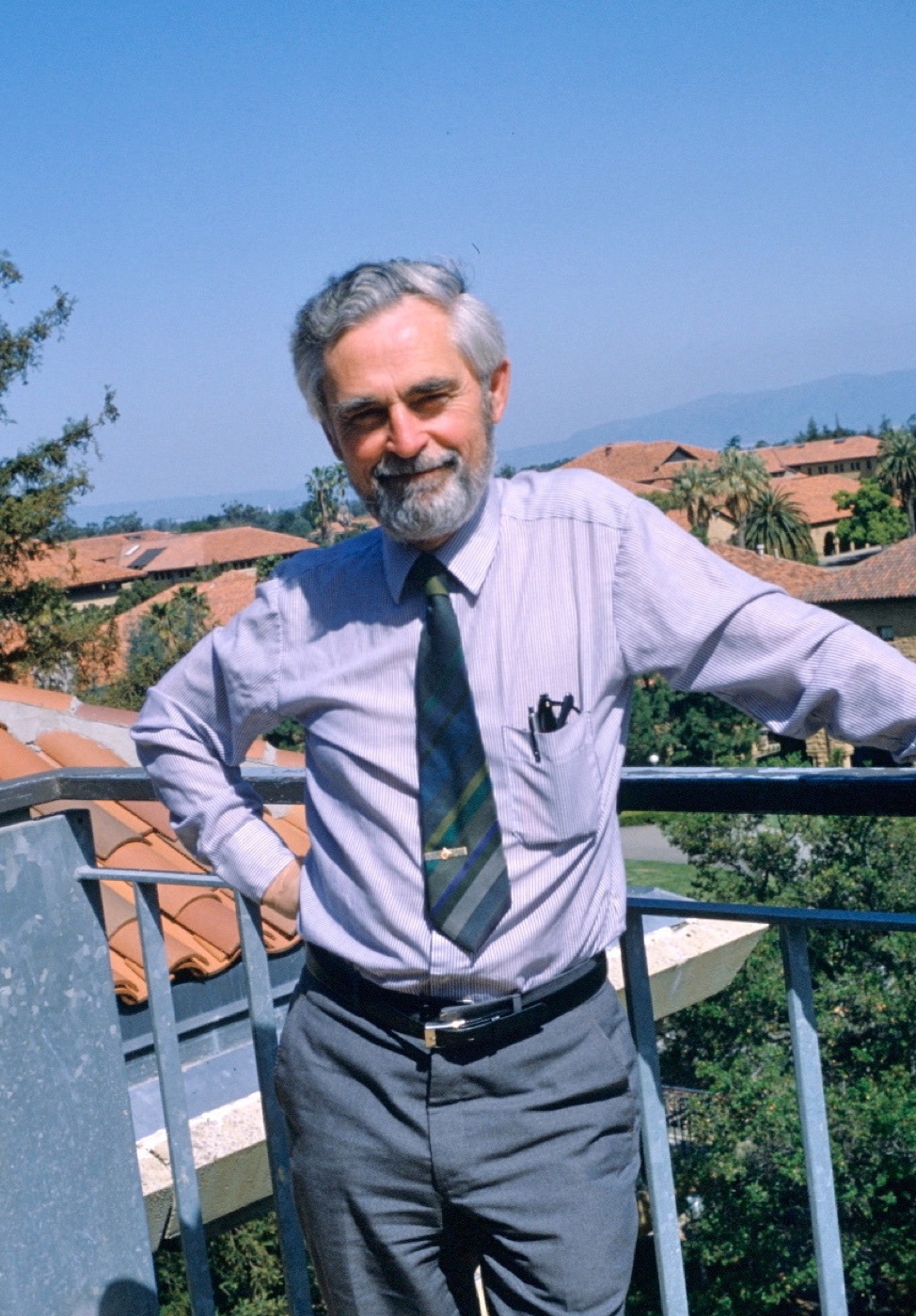 Grey-bearded man in a striped shirt and tie leaning on a rooftop railing with tiled Stanford roofs behind him