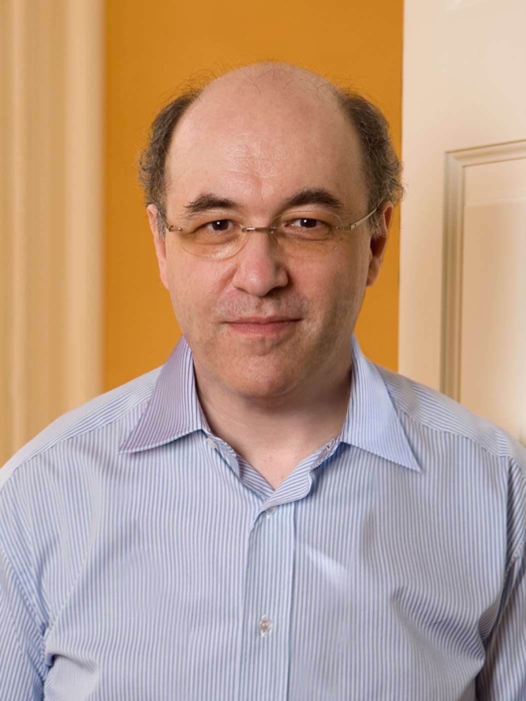 Portrait of a bald man in glasses and a blue striped shirt in front of an orange wall